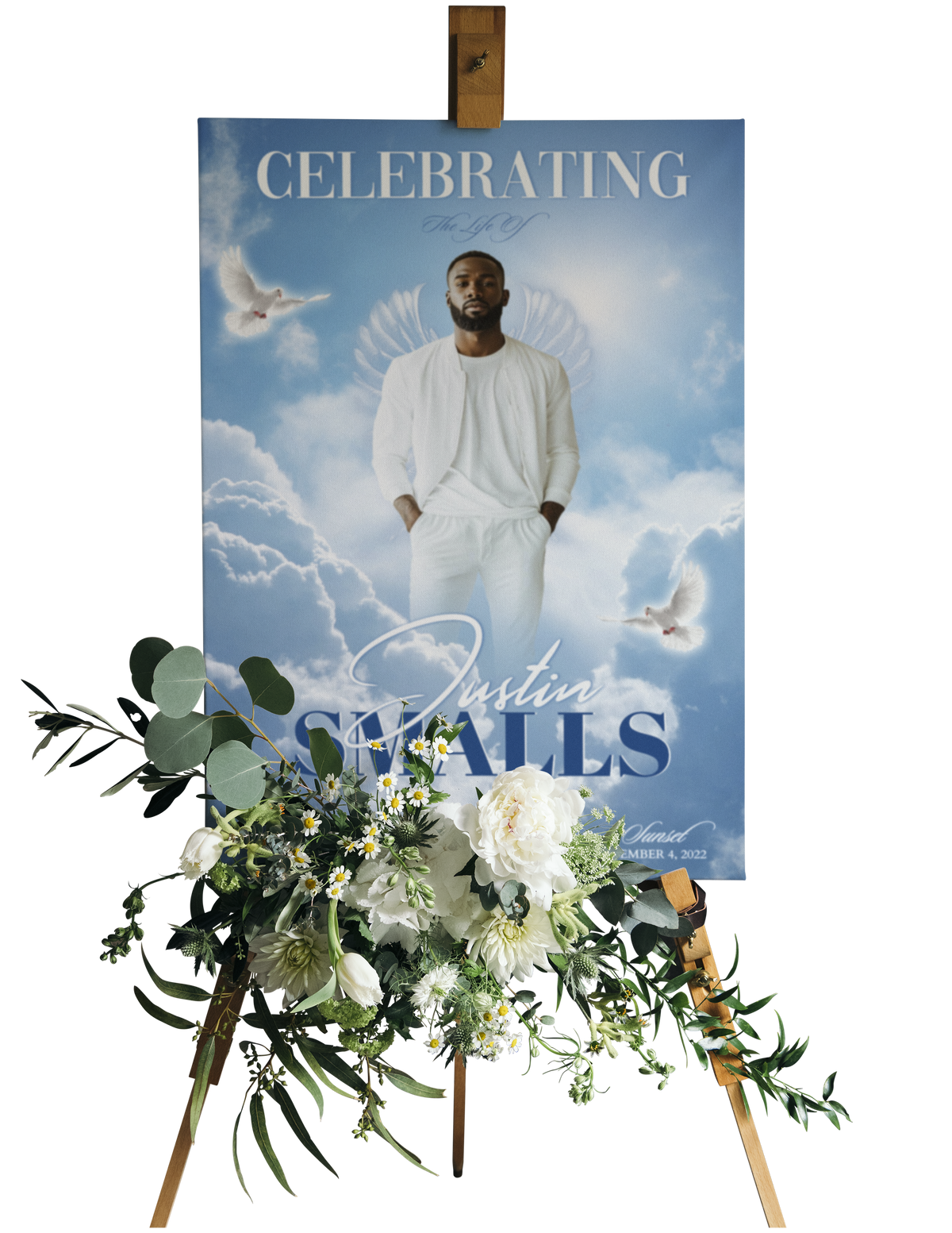 A personalized memorial or celebration poster displayed on a wooden easel, decorated with a floral arrangement in the foreground. The poster features a portrait of a man in a white outfit against a blue sky with clouds and doves, with the text 'CELEBRATING' and 'with Justin SMALLS'.